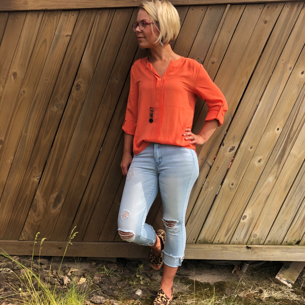 A woman wearing an orange chiffon v-neck top with adjustable roll-up sleeves and a single button detail, paired with blue jeans. The setting suggests a casual outdoor environment.